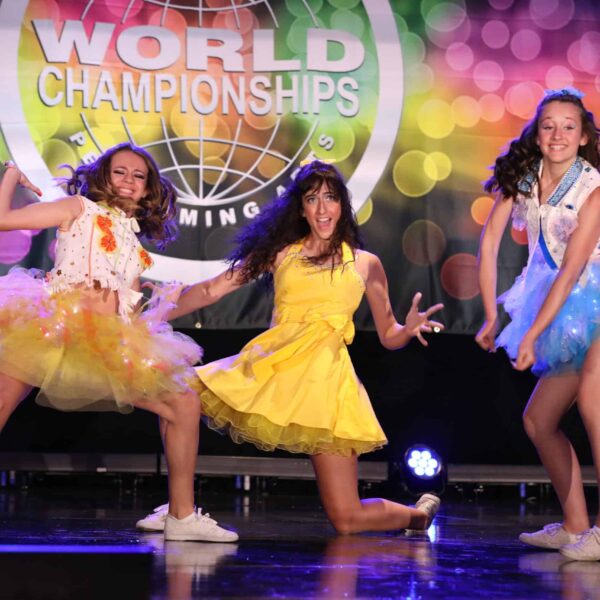 Trois jeunes danseuses en tutus colorés devant l'affiche World Championships lors d'un spectacle dynamique.